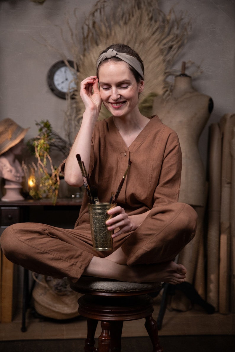 A smiling girl is sitting on a round vintage chair, holding a brass glass with paintbrushes. She is also wearing the 'Artist' jumpsuit from Knock Knock Linen