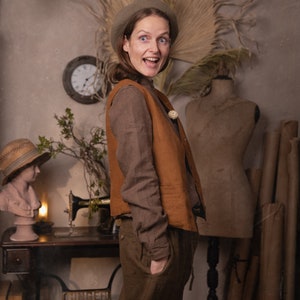 Side view of a model wearing a brown linen shirt under a rust-colored linen vest with buttons, paired with olive linen pants and a beige beret, hands in pocket, standing near a vintage sewing table, knockknocklinen.