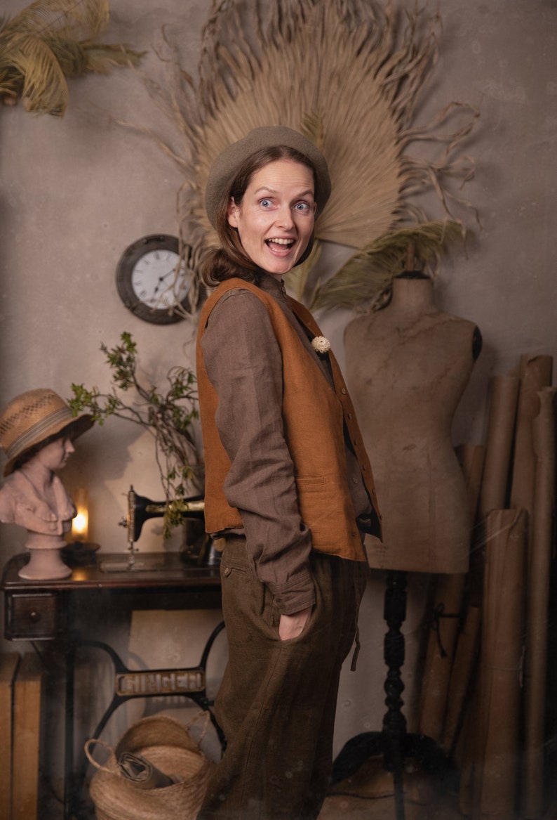Side view of a model wearing a brown linen shirt under a rust-colored linen vest with buttons, paired with olive linen pants and a beige beret, hands in pocket, standing near a vintage sewing table, knockknocklinen.