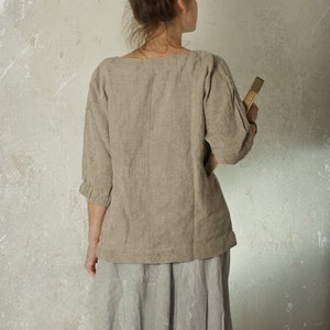 Person facing away from the camera, wearing a loose natural herringbone linen blouse made from flax, with three-quarter sleeves, paired with a light grey knee-length skirt. Hair in a messy bun, holding a book. Shown against textured wall. Linen blouse, loose linen top.