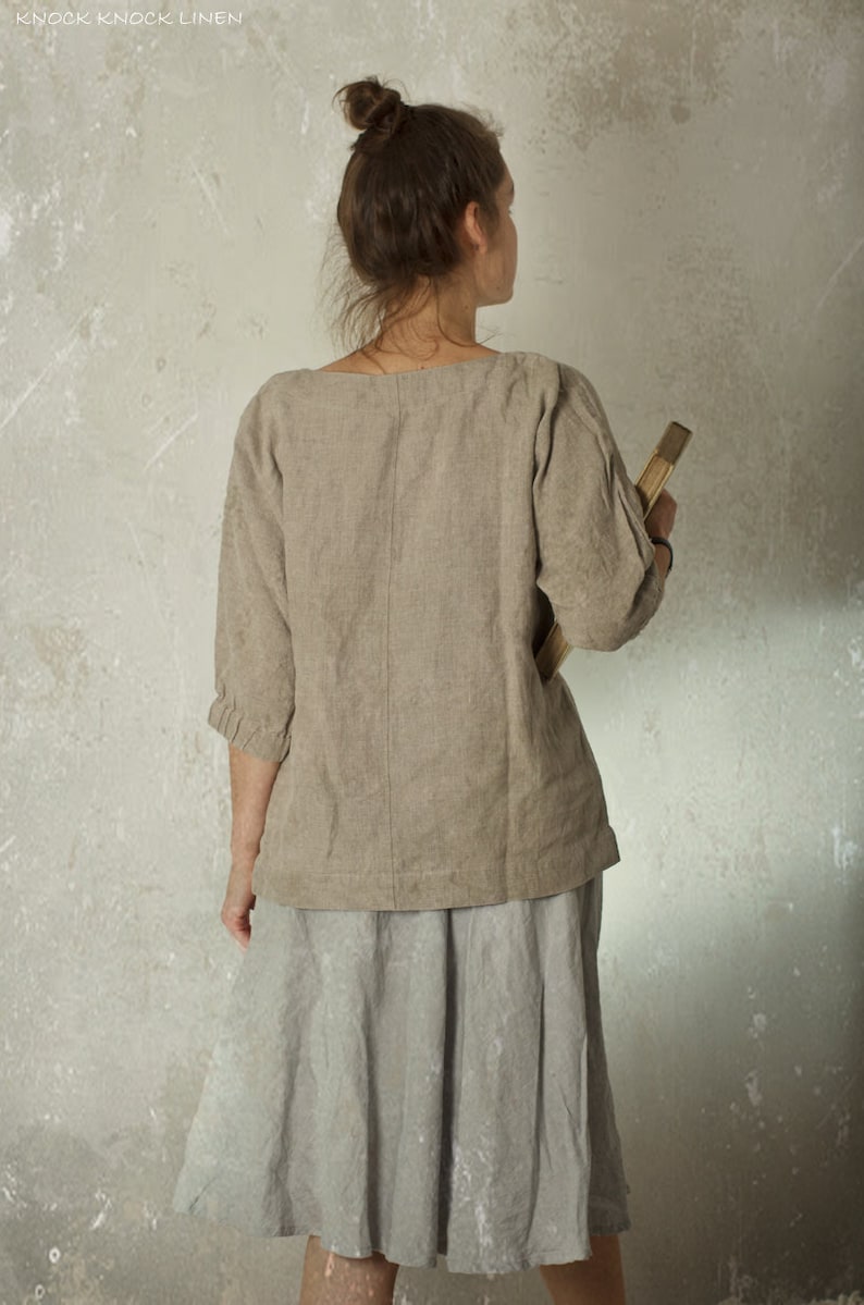 Person facing away from the camera, wearing a loose natural herringbone linen blouse made from flax, with three-quarter sleeves, paired with a light grey knee-length skirt. Hair in a messy bun, holding a book. Shown against textured wall. Linen blouse, loose linen top.