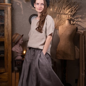 Model indoors wearing gray linen maxi skirt with elastic waistband, drawstring waist, and side pockets; loose fit and floor-length. Paired with light gray linen top. Decorative props include vintage dress form, clock, and dried plants. Keywords: peasant skirt, linen summer skirt.