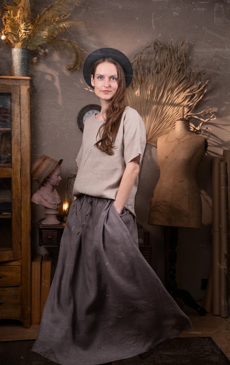 Model indoors wearing gray linen maxi skirt with elastic waistband, drawstring waist, and side pockets; loose fit and floor-length. Paired with light gray linen top. Decorative props include vintage dress form, clock, and dried plants. Keywords: peasant skirt, linen summer skirt.