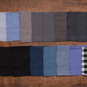 Linen fabric samples in pale gray, light blue, taupe, charcoal, navy, and black displayed in a flat lay close-up, set in two rows on a brown wooden surface, with zigzag-cut edges, rectangular pieces, a striped swatch, and a checked swatch.