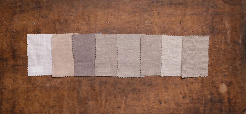 KnockKnockLinen linen fabric swatches—8 neutral samples (white, beige, taupe, gray) in plain weave and subtle herringbone, pinked edges, arranged in one row; flat lay on a rustic wooden surface.