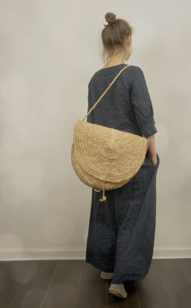 Back view of one model wearing a dark navy blue linen maxi dress with 3/4 sleeves and side pockets, kaftan style and loose fitting, standing indoors on dark wooden floor, styled with a large woven straw shoulder bag.