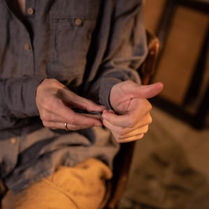 Close-up of a seated model in a gray linen button-up shirt with light brown buttons and chest pocket, hands in focus against a warm blurred background, knockknocklinen.