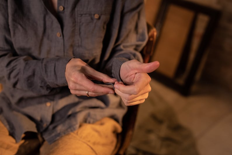 Close-up of a seated model in a gray linen button-up shirt with light brown buttons and chest pocket, hands in focus against a warm blurred background, knockknocklinen.