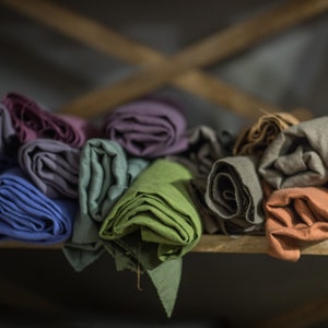 Puede incluir: Una pila de tela enrollada en varios colores, incluyendo verde, morado, gris y marrón. La tela está dispuesta en un estante de madera.