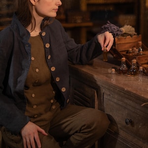 SPARROW linen vest in brown, shown on a seated model in a front three-quarter mid-length photo, with a round neckline, two rows of light buttons, and an angled overlapping front, layered under a dark blue jacket, Knock Knock Linen.