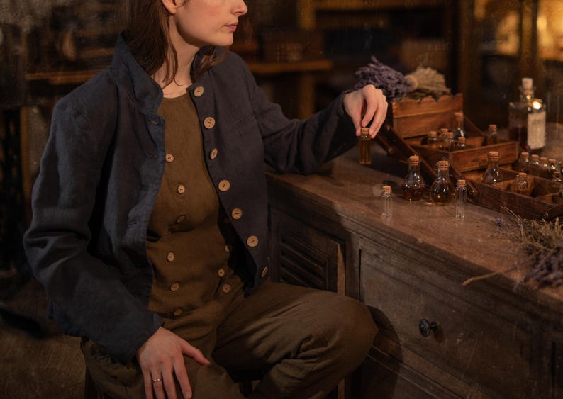 SPARROW linen vest in brown, shown on a seated model in a front three-quarter mid-length photo, with a round neckline, two rows of light buttons, and an angled overlapping front, layered under a dark blue jacket, Knock Knock Linen.