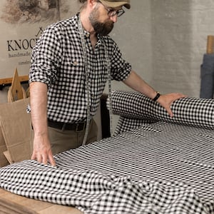Three-quarter, waist-up view of black-and-white checkered linen fabric spread on a wooden worktable, with a matching fabric roll at right, bearded man in a checked shirt and beige cap holding the cloth, and a measuring tape around his neck.