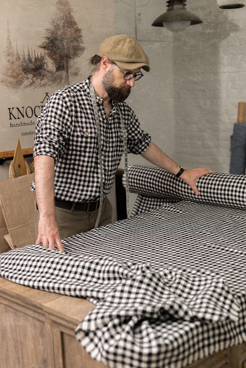 Three-quarter, waist-up view of black-and-white checkered linen fabric spread on a wooden worktable, with a matching fabric roll at right, bearded man in a checked shirt and beige cap holding the cloth, and a measuring tape around his neck.