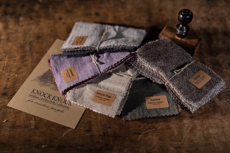 KnockKnockLinen all-in-one fabric samples—several soft linen swatch bundles in gray, lavender, natural flax, cactus green and textured brown, tied with string and labeled; flat lay on a wooden table with a branded card.