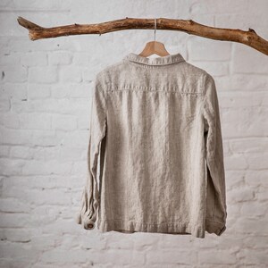Natural Flax linen shirt with long sleeves, buttoned cuffs, peter pan collar, and back yoke detail, hanging on wooden hanger from branch against white brick wall. Loose linen jacket for women, suitable as workwear or summer jacket.