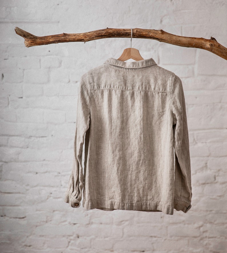 Natural Flax linen shirt with long sleeves, buttoned cuffs, peter pan collar, and back yoke detail, hanging on wooden hanger from branch against white brick wall. Loose linen jacket for women, suitable as workwear or summer jacket.