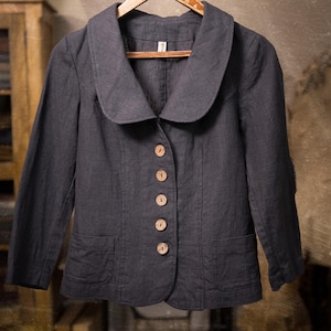 Charcoal linen jacket shown hanging on a wooden hanger in a front full-length view, with a wide rounded collar, five light wooden buttons, long sleeves, two front patch pockets, and a slightly curved hem.