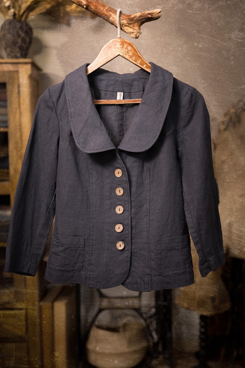 Charcoal linen jacket shown hanging on a wooden hanger in a front full-length view, with a wide rounded collar, five light wooden buttons, long sleeves, two front patch pockets, and a slightly curved hem.