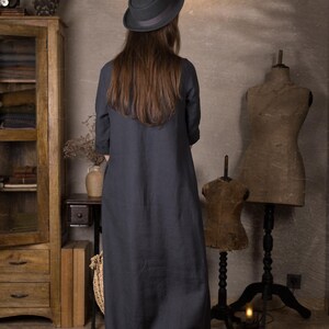 Rear view of a loose dark blue long linen dress with 3/4 sleeves shown on a model wearing a black hat and brown shoes, standing in a neutral vintage-style room.