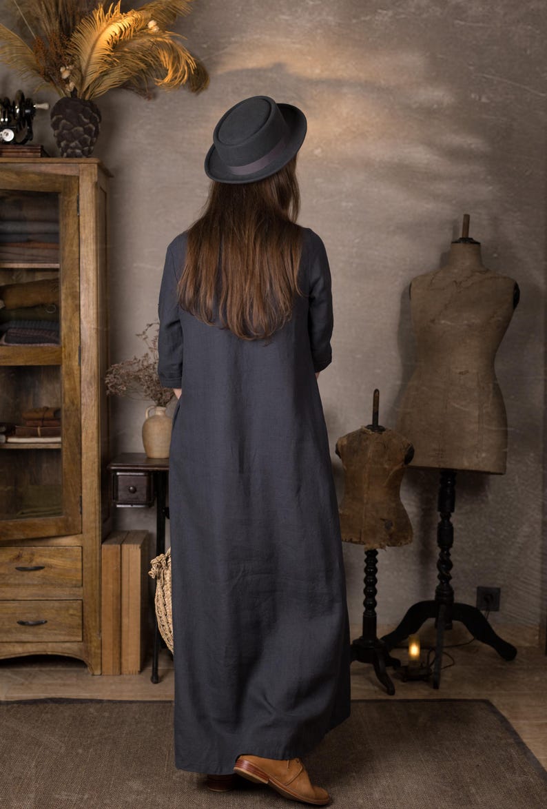 Rear view of a loose dark blue long linen dress with 3/4 sleeves shown on a model wearing a black hat and brown shoes, standing in a neutral vintage-style room.