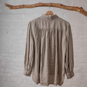 Natural taupe linen blouse CLAIRE displayed on a wooden hanger in a full back view, with a folded collar, horizontal back yoke, gathered fabric below the yoke, long sleeves, buttoned cuffs, and a curved hem.