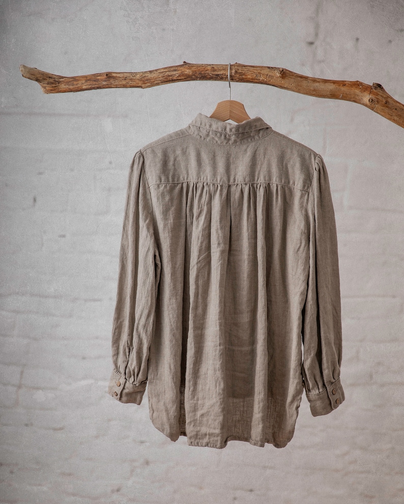 Natural taupe linen blouse CLAIRE displayed on a wooden hanger in a full back view, with a folded collar, horizontal back yoke, gathered fabric below the yoke, long sleeves, buttoned cuffs, and a curved hem.