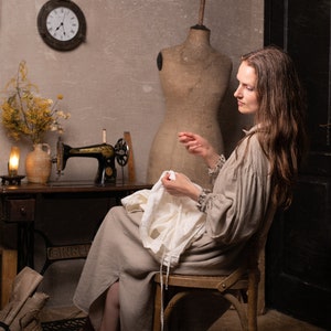 Light beige linen dress with long sleeves and gathered cuffs, seated on a wooden chair holding white fabric in a sewing room with wall clock and mannequin.