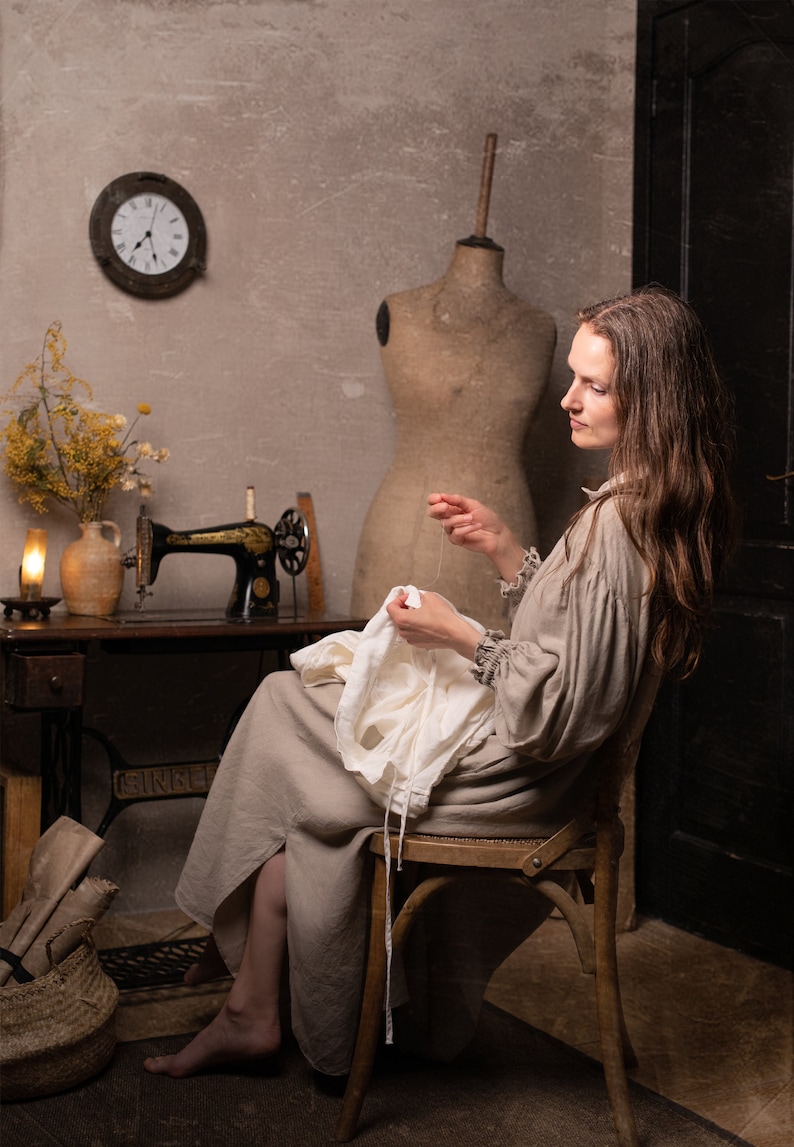 Light beige linen dress with long sleeves and gathered cuffs, seated on a wooden chair holding white fabric in a sewing room with wall clock and mannequin.