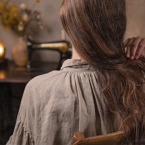 Close-up of a model from behind wearing one natural linen dress with gathered back neckline, seated on a wooden chair indoors.