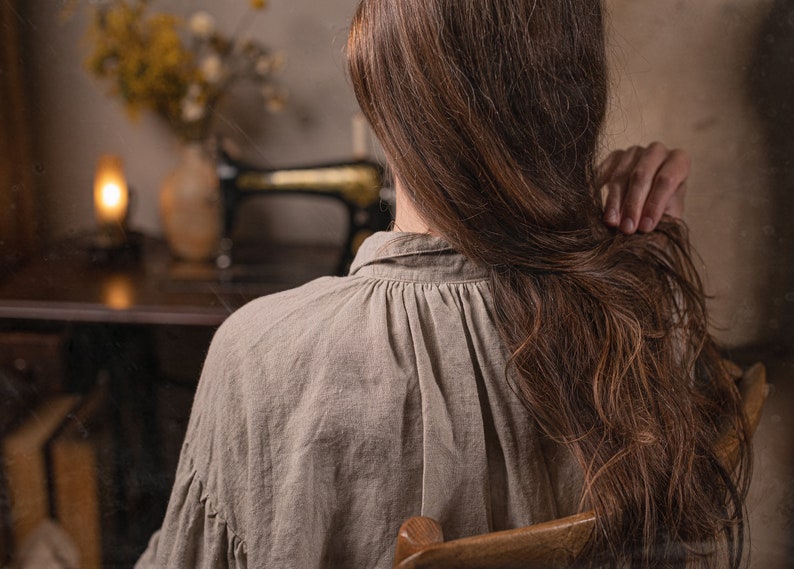 Close-up of a model from behind wearing one natural linen dress with gathered back neckline, seated on a wooden chair indoors.