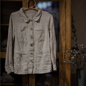 Natural Flax linen shirt FACTORY with long sleeves, button front, peter pan collar, and two front pockets, hanging on wooden hanger in front of glass cabinet with folded clothes. Loose linen jacket for women, styled as workwear or summer jacket.