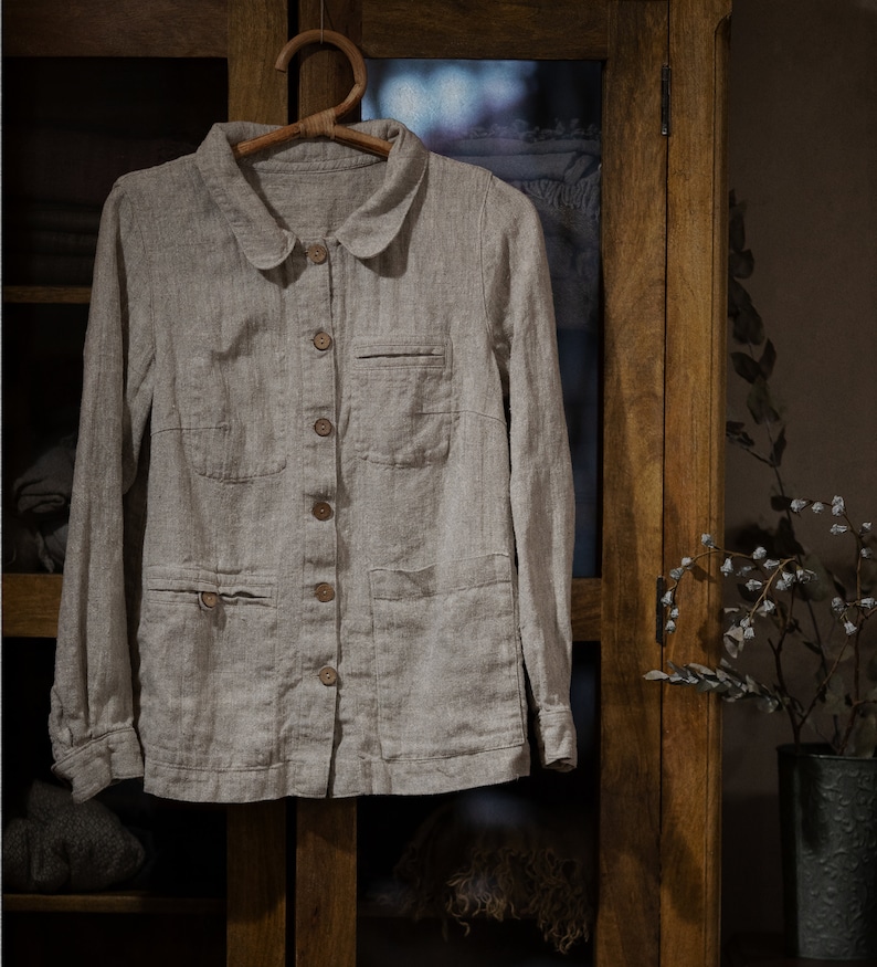 Natural Flax linen shirt FACTORY with long sleeves, button front, peter pan collar, and two front pockets, hanging on wooden hanger in front of glass cabinet with folded clothes. Loose linen jacket for women, styled as workwear or summer jacket.