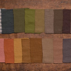 Linen fabric samples in muted gray, green, beige, brown, pink, and blue-gray colors displayed in a flat lay detail view, set in two neat rows on dark wood, with zigzag-cut edges, plain weaves, and one small check swatch.