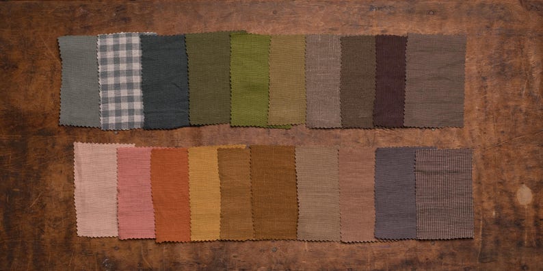 Linen fabric samples in muted gray, green, beige, brown, pink, and blue-gray colors displayed in a flat lay detail view, set in two neat rows on dark wood, with zigzag-cut edges, plain weaves, and one small check swatch.