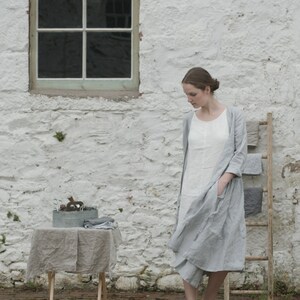 A person wearing a light grey linen dress and grey shoes stands in front of a white stone wall with a window. To the left, a small table covered with a grey cloth holds a metal container with various items. Behind the person, a wooden ladder displays folded grey and white linens. The brick-paved ground adds texture to the scene.