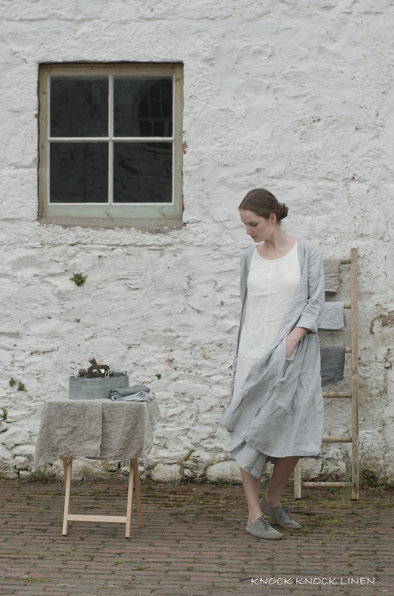 A person wearing a light grey linen dress and grey shoes stands in front of a white stone wall with a window. To the left, a small table covered with a grey cloth holds a metal container with various items. Behind the person, a wooden ladder displays folded grey and white linens. The brick-paved ground adds texture to the scene.