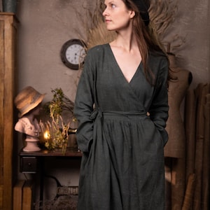 A person wears a dark green wrap dress with long sleeves, tied at the waist, and a black hat. The vintage-style room features a mannequin, a straw-hat-wearing bust, a clock, and dried plants.