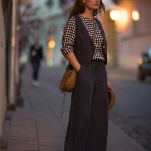 Charcoal  linen wide-leg palazzo pants on model, full-length three-quarter view; high waist with smooth front, one hand in side pocket, loose drape with vertical folds, floor-length hem over sandals; blurred streetlights behind.