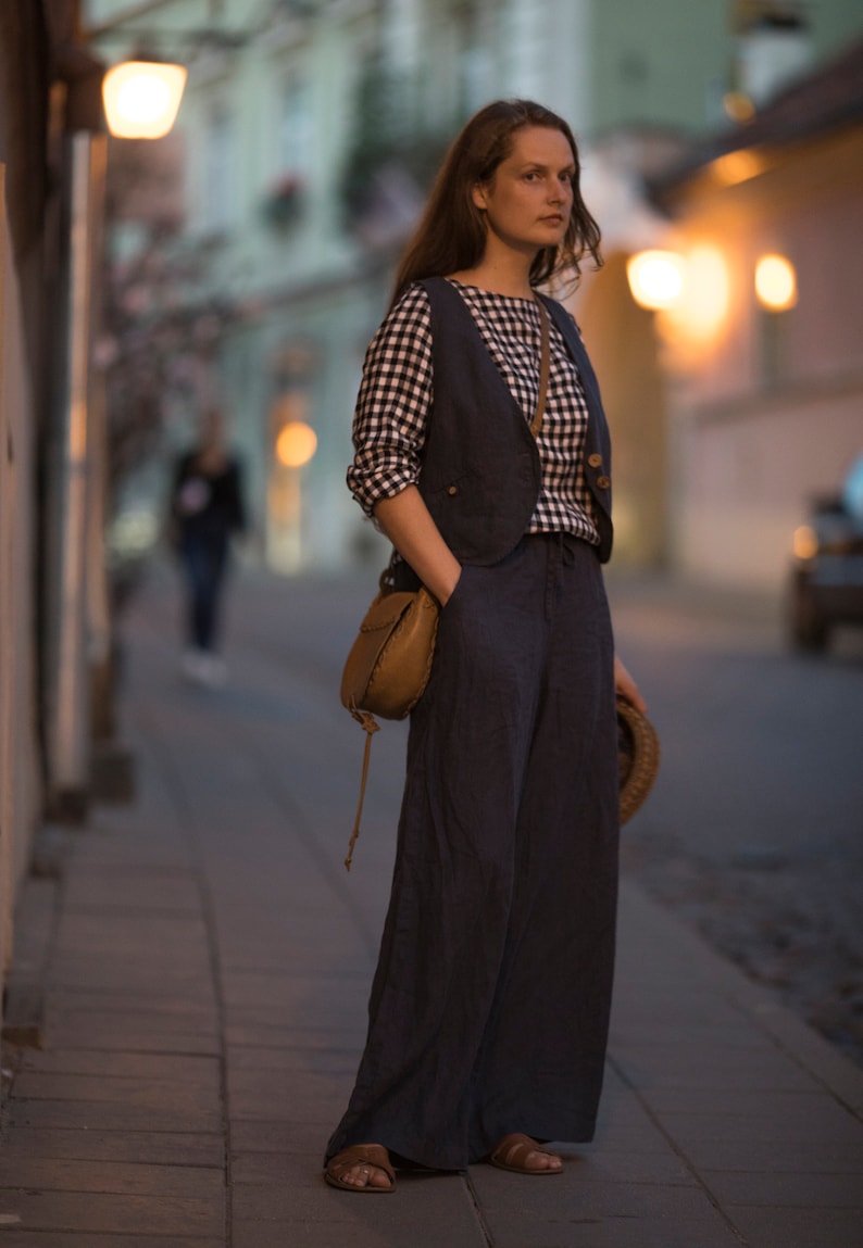 Charcoal  linen wide-leg palazzo pants on model, full-length three-quarter view; high waist with smooth front, one hand in side pocket, loose drape with vertical folds, floor-length hem over sandals; blurred streetlights behind.