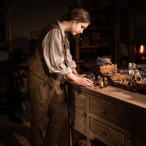 SPARROW linen vest in brown, displayed on a model in a side three-quarter full-length view, with a sleeveless shape, a long side line, worn over a light gray blouse with puffed sleeves and matching brown trousers, beside glass bottles and lavender, Knock Knock Linen.