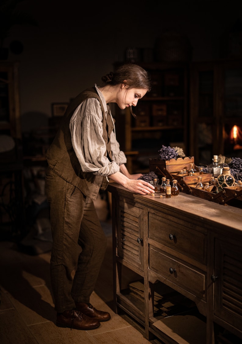 SPARROW linen vest in brown, displayed on a model in a side three-quarter full-length view, with a sleeveless shape, a long side line, worn over a light gray blouse with puffed sleeves and matching brown trousers, beside glass bottles and lavender, Knock Knock Linen.