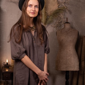 Dark taupe linen NOMAD dress on a model in a front three-quarter crop, with gathered neckline, short button placket, puff sleeves, rolled cuffs, side slit, and loose straight shape.