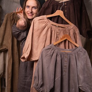 Person wearing one dark-colored linen boho dress with short sleeves and pockets, styled with black hat. Standing behind hanging dresses in brown, beige, and gray linen on wooden hangers. Rustic indoor setting with dried plants. Linen summer dress display.