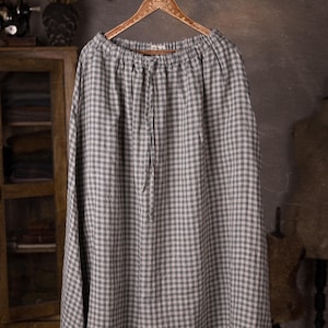 Gray and white gingham linen skirt shown full-length on an ornate wooden hanger, front view, ruched waistband with drawstring channel, smooth skirt panels with no visible pockets, wide hemline, cabinet and dress forms behind.