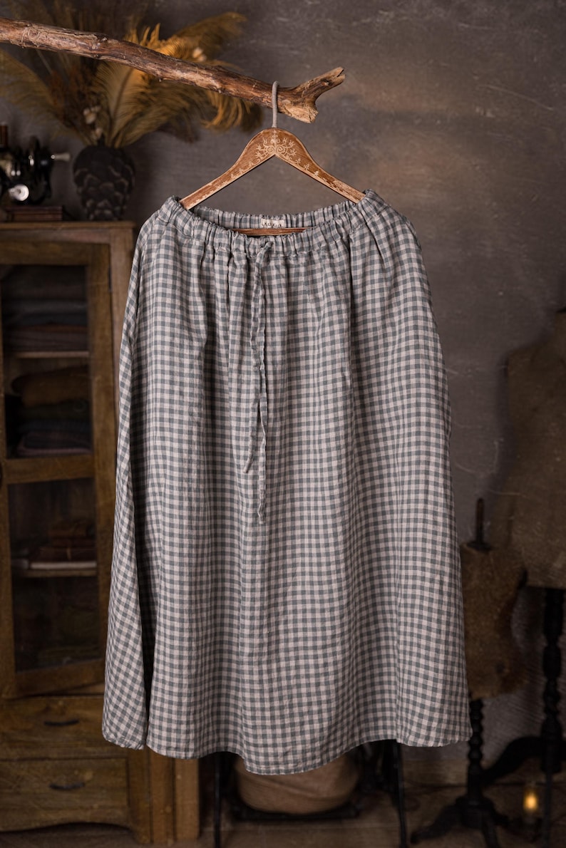 Gray and white gingham linen skirt shown full-length on an ornate wooden hanger, front view, ruched waistband with drawstring channel, smooth skirt panels with no visible pockets, wide hemline, cabinet and dress forms behind.