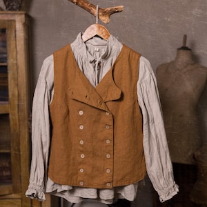 SPARROW linen vest in brown, shown on a hanger in a front-view full-length photo, with a sleeveless shape, double rows of light buttons, an offset front panel, and a straight lower edge, over a light gray blouse with long gathered sleeves, Knock Knock Linen.
