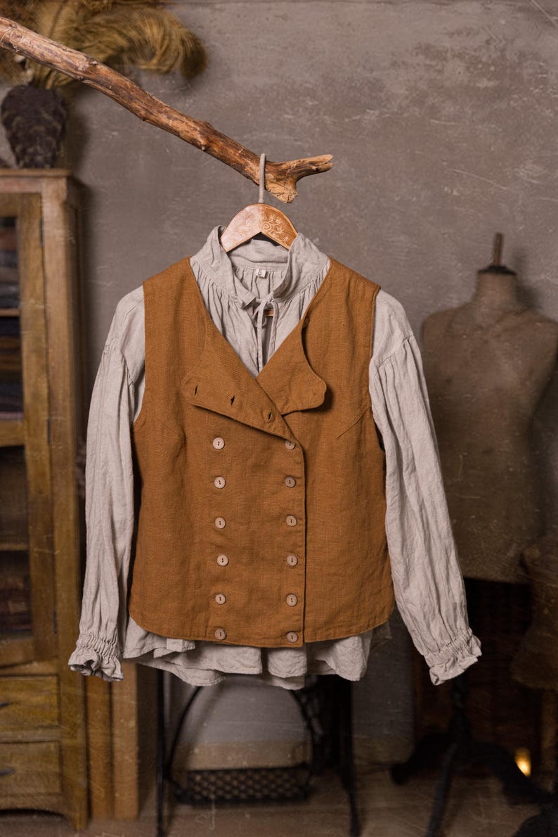 SPARROW linen vest in brown, shown on a hanger in a front-view full-length photo, with a sleeveless shape, double rows of light buttons, an offset front panel, and a straight lower edge, over a light gray blouse with long gathered sleeves, Knock Knock Linen.