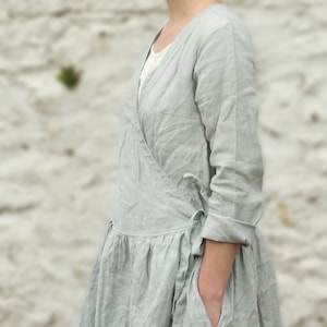 A model wearing a light gray, long-sleeve, mid-calf linen wrap dress with a tie at the waist and pockets. The dress has a natural, slightly wrinkled texture and is styled in a relaxed, cottagecore fashion. The background is a blurred, neutral outdoor setting.