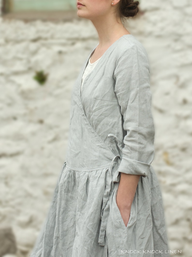 A model wearing a light gray, long-sleeve, mid-calf linen wrap dress with a tie at the waist and pockets. The dress has a natural, slightly wrinkled texture and is styled in a relaxed, cottagecore fashion. The background is a blurred, neutral outdoor setting.