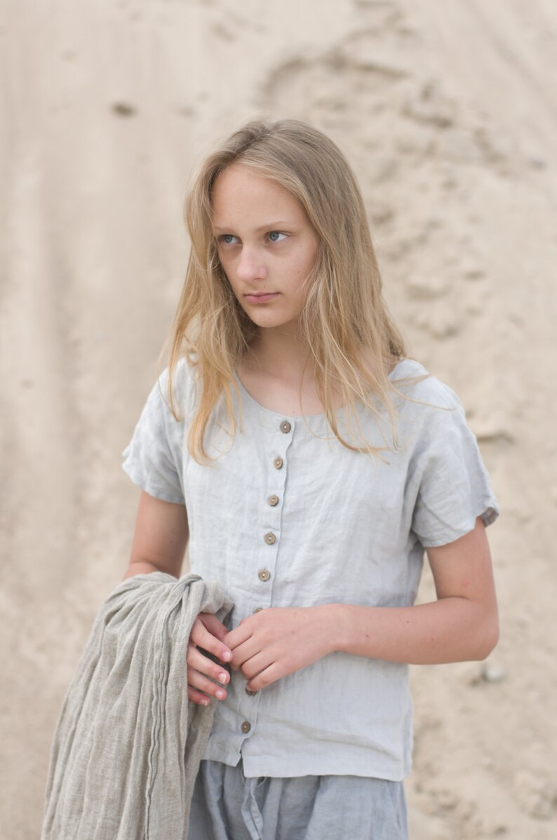 Woman wearing a dusty sky linen buttoned top with short sleeves and round neckline. Five buttons run down the front. She holds a matching linen cloth. Photographed outdoors on sandy beach. Linen blouse, buttoned shirt, loose linen top.